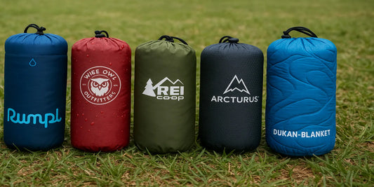 Five waterproof camping blankets in branded carrying cases, displayed on grass, featuring Rumpl, Wise Owl, REI, Arcturus, and Dukan.